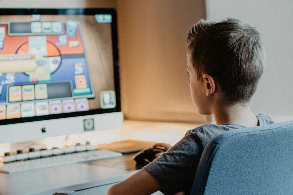 An over-the-shoulder view of a young person seated at a desk, looking at a computer monitor. The individual is playing a digital trading card game, with various cards and gameplay elements visible on the screen. The setting is a brightly lit room with a keyboard and mouse on the desk.